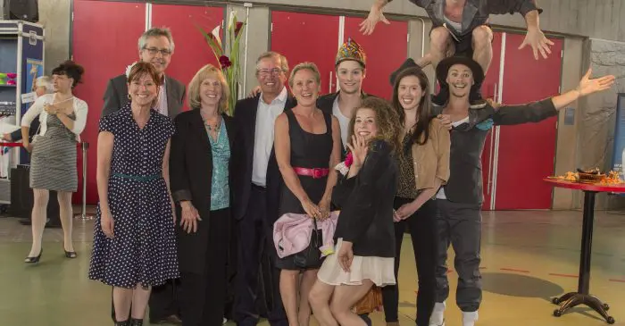 Un groupe de personnes pose ensemble à l'intérieur lors de la soirée-bénéfice 2015 : plus de 171 000$ de récolté, certaines habillées de manière formelle et d'autres en costumes de cirque. Deux artistes enjoués se distinguent, l'un perché sur les épaules de l'autre.
