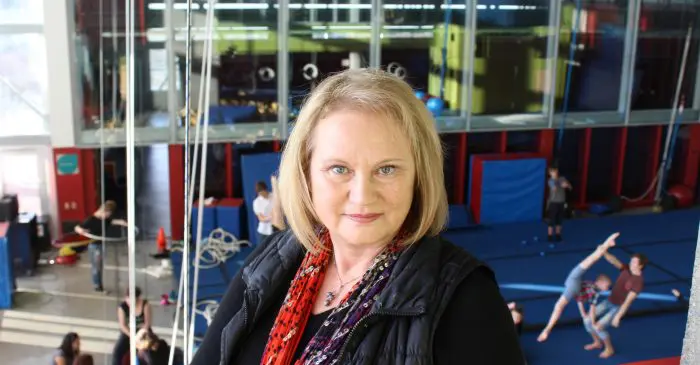 Une femme aux cheveux blonds, portant un gilet noir et une écharpe rouge, se tient à l'intérieur devant de grandes fenêtres. Derrière elle, des personnes pratiquent la gymnastique sur des tapis bleus dans un gymnase lumineux où Daniela Arendasova préside le jury du Festival Mondial du Cirque de Demain.