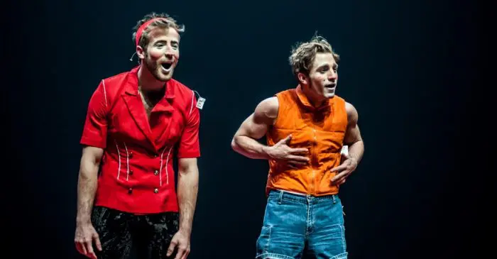Deux artistes masculins se tiennent sur scène sous un éclairage dramatique lors du 37e Festival mondial du Cirque de Demain, avec des visages expressifs en pleine représentation. L'un porte une chemise rouge et un pantalon noir, l'autre un haut orange sans manches et un short en jean.