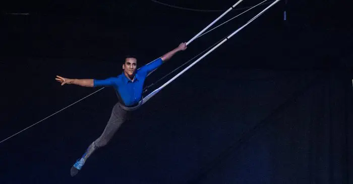 Un acrobate de cirque en chemise bleue et pantalon gris se balance gracieusement sur des cordes au-dessus d'une scène sombre, un bras tendu vers l'extérieur dans une pose dramatique - rappelant les six diplômé-e-s qui ont décroché les plus grands honneurs au Festival Mondial du Cirque de Demain.