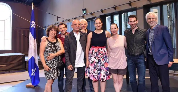 Un groupe de huit personnes se tient en souriant à l'intérieur, à côté d'un drapeau bleu et blanc avec un symbole de fleur de lys, célébrant un octroi de 1 million de dollars pour développer la pratique du cirque. Une scène et une grande fenêtre apparaissent à l'arrière-plan.