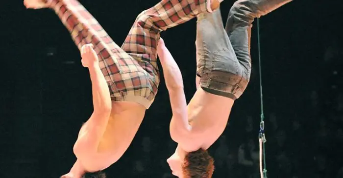 Deux acrobates torse nu effectuent des sauts synchronisés sur scène, vêtus de pantalons écossais et gris, sous les lumières de la scène et sous les yeux du public - un spectacle conçu alors que cinq concepteur-rice-s aux parcours artistiques riches et variés sont de passage à l'ECN.