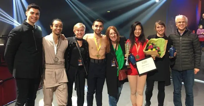 Un groupe de huit personnes pose ensemble sur une scène, certaines portant des médailles - un triplé pour les diplômé-e-s de l'ECN au Festival Mondial du Cirque de Demain. Une personne tient un certificat et des fleurs sous des lumières vives et des drapeaux internationaux.
