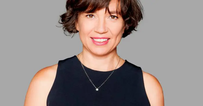 Une femme aux cheveux bruns courts et au haut noir sans manches sourit devant un fond gris uni, célébrant la nomination de Mme Estelle Clareton à titre de directrice de la création. Elle porte un collier simple.