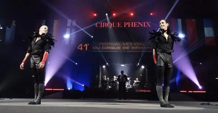 Deux artistes en noir avec des épaules en plumes se tiennent sur la scène du Cirque Phénix, sous les projecteurs, avec des musiciens et des drapeaux internationaux derrière eux - lauréats de la médaille d'or au terme de la 41e édition du Festival Mondial du Cirque de Demain.