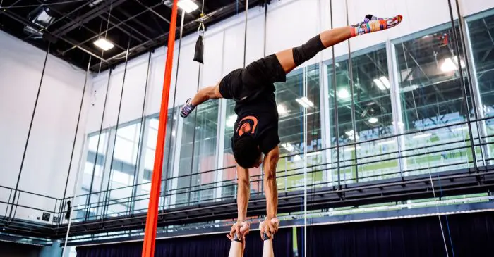 Une personne se tient en équilibre sur les mains d'une autre, les jambes écartées, à l'intérieur d'un gymnase doté de grandes fenêtres, de soies aériennes et de hauts plafonds - un espace qui valorise la diversité et l'inclusion.