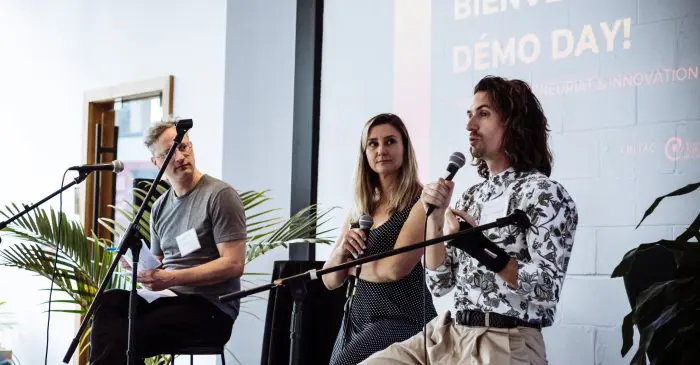 Trois personnes sont assises sur des tabourets et tiennent des microphones. Elles prennent la parole lors d'un événement en intérieur. Derrière eux, un écran affiche "Bienvenue au Démo Day !" et les détails de l'événement "Le CRITAC lance son lab en entrepreneuriat et innovation". Deux grandes plantes en pot encadrent la scène.