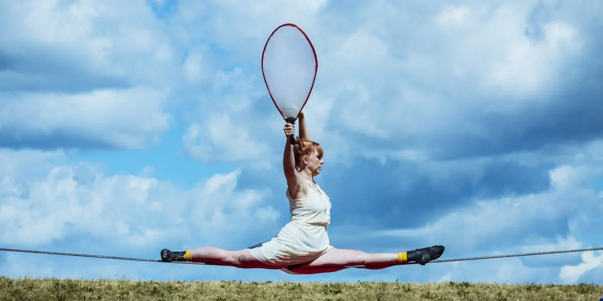 Une femme en tenue blanche fait un grand écart sur une corde raide en plein air, tenant une raquette de tennis géante, dans le cadre du spectacle extérieur 2021 entre-temps des étudiant-e-s de l'ENC, avec un terrain herbeux et un ciel partiellement nuageux en arrière-plan.