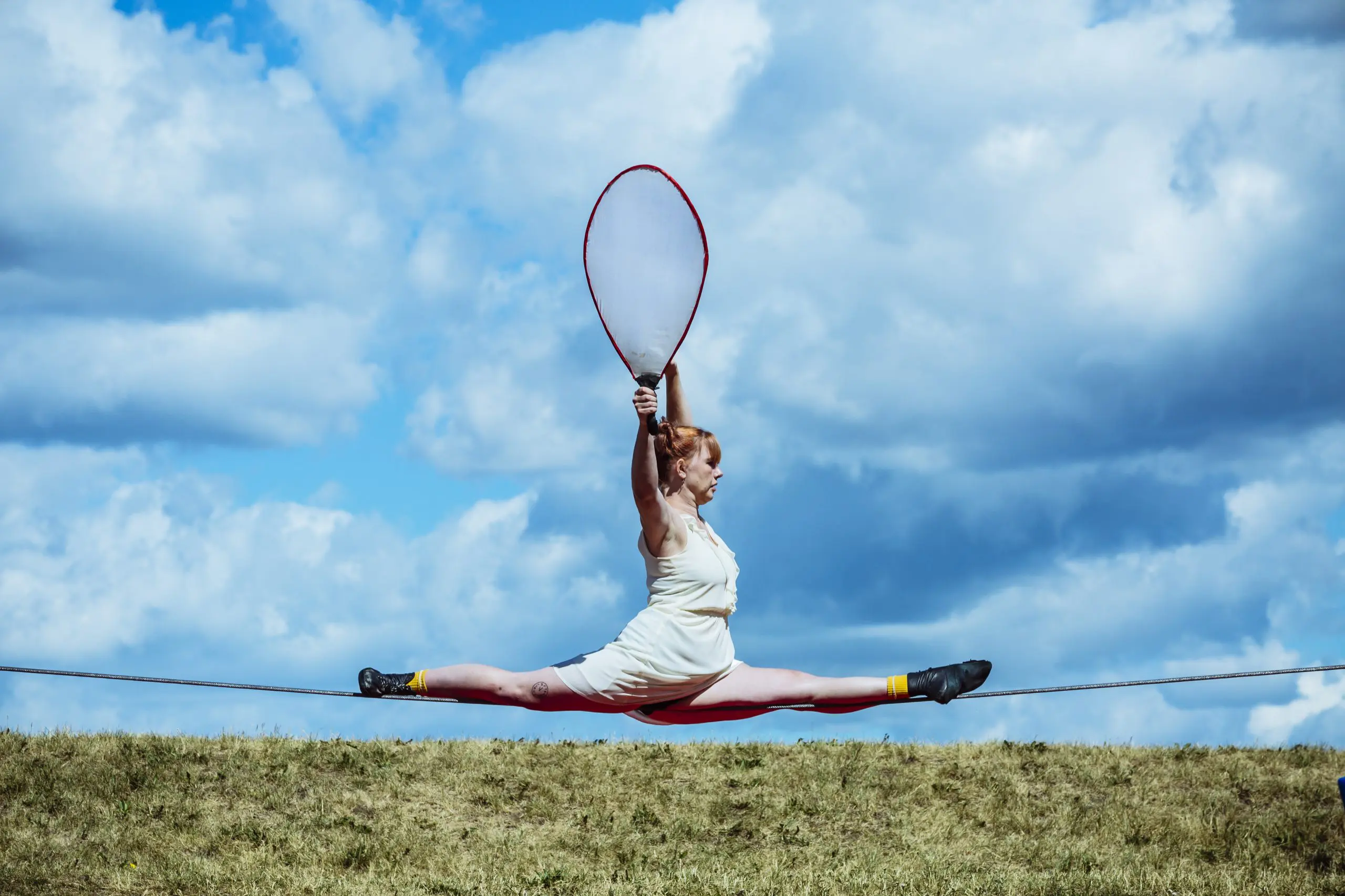 Une femme en tenue blanche fait un grand écart sur une corde raide en plein air, tenant une raquette de tennis géante, dans le cadre du spectacle extérieur 2021 entre-temps des étudiant-e-s de l'ENC, avec un terrain herbeux et un ciel partiellement nuageux en arrière-plan.