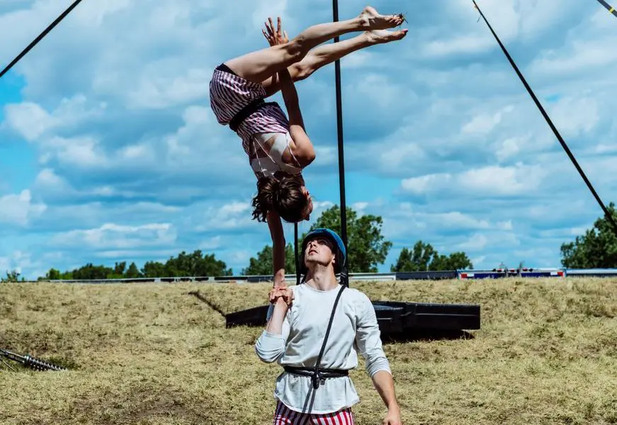 Un homme en short rayé fait tenir une femme à l'envers sur une main lors d'un spectacle extérieur 2021 entre-temps des étudiant-e-s de l'ENC, sur fond de terrain herbeux et de ciel nuageux.