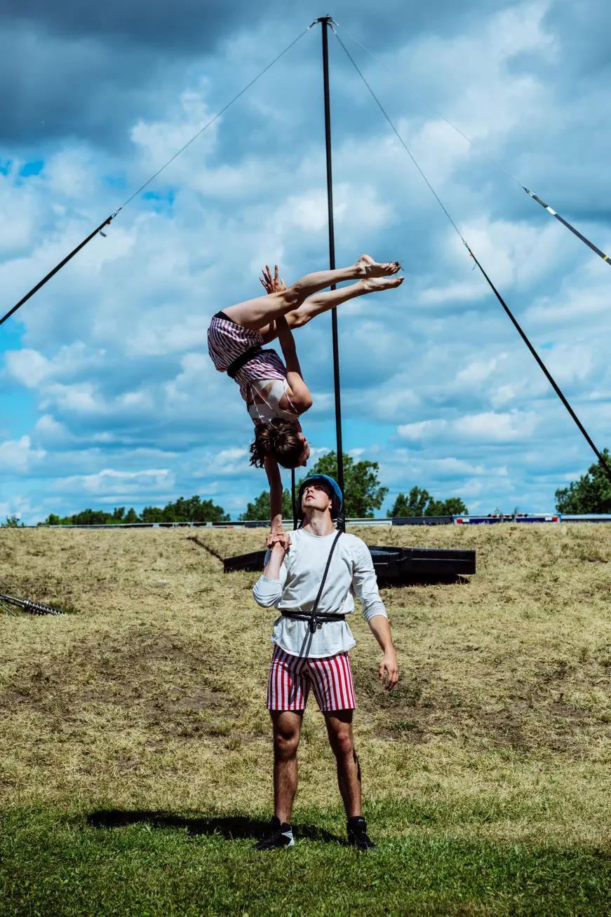 Un homme en short rayé fait tenir une femme à l'envers sur une main lors d'un spectacle extérieur 2021 entre-temps des étudiant-e-s de l'ENC, sur fond de terrain herbeux et de ciel nuageux.