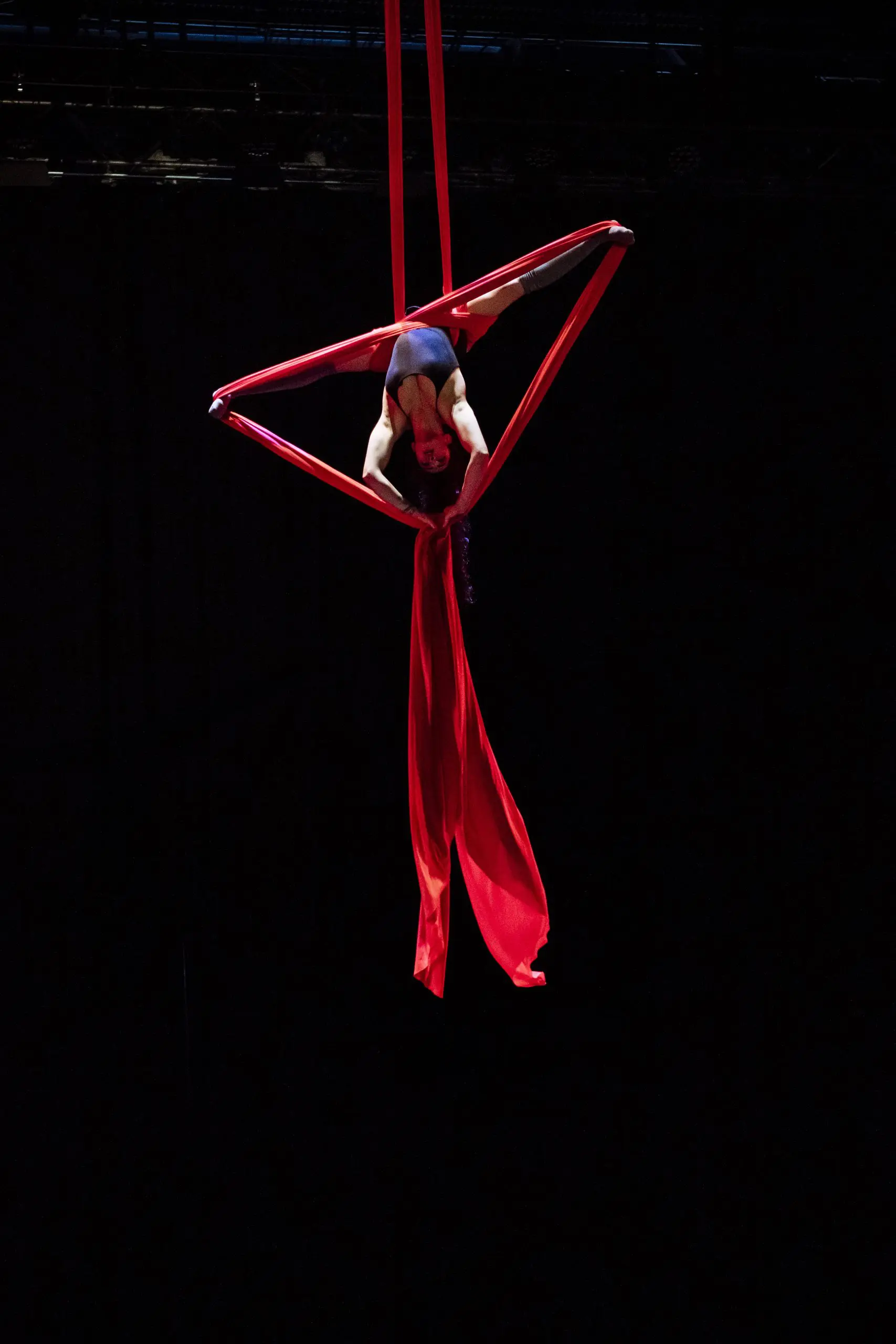 L'artiste aérienne Leela Masuret prend la pose à l'envers sur un tissu de soie rouge, suspendue dans les airs sur un fond sombre.