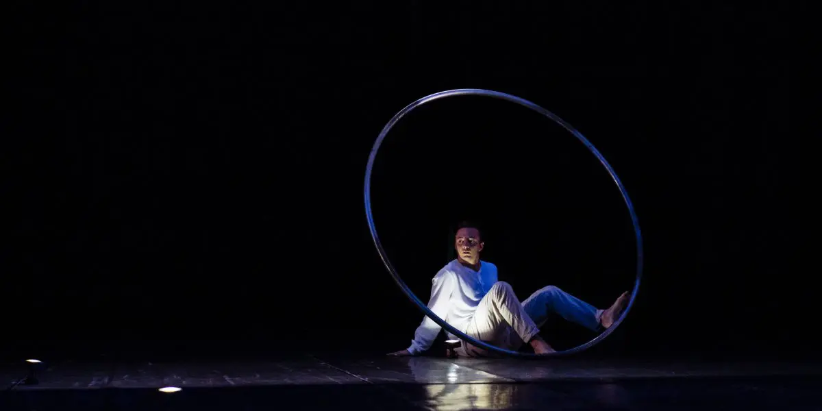 Un artiste vêtu de blanc est assis sur le sol à l'intérieur d'un grand cerceau métallique, éclairé par un projecteur sur une scène sombre - une scène captivante chorégraphiée par Robert Cookson.