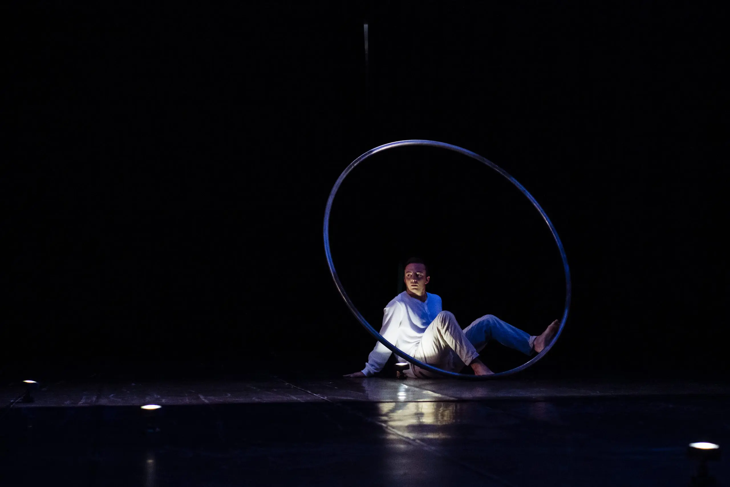 Un artiste vêtu de blanc est assis sur le sol à l'intérieur d'un grand cerceau métallique, éclairé par un projecteur sur une scène sombre - une scène captivante chorégraphiée par Robert Cookson.