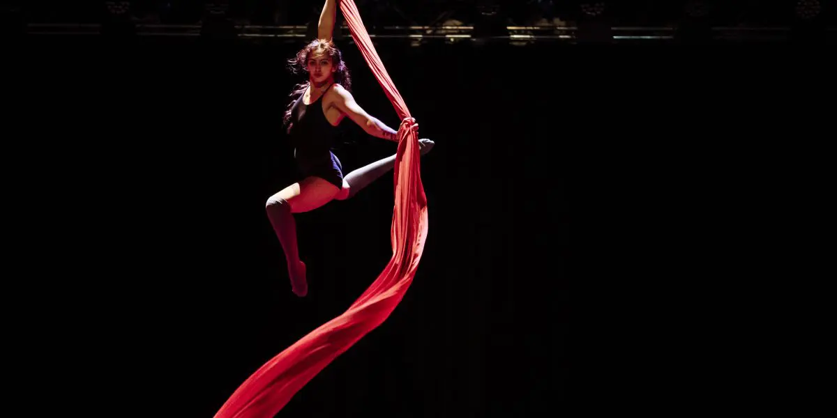 L'artiste aérienne Leela Masuret est suspendue dans les airs sur un tissu de soie rouge, un bras tenant le tissu et les jambes étendues avec grâce sur un fond de scène sombre.