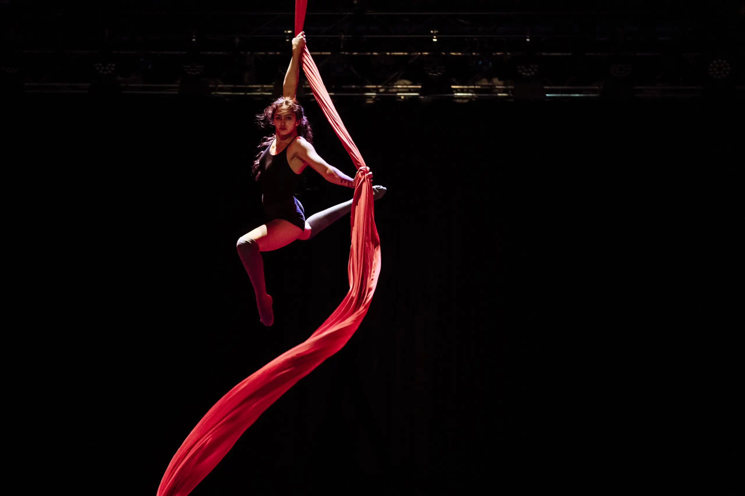 L'artiste aérienne Leela Masuret est suspendue dans les airs sur un tissu de soie rouge, un bras tenant le tissu et les jambes étendues avec grâce sur un fond de scène sombre.