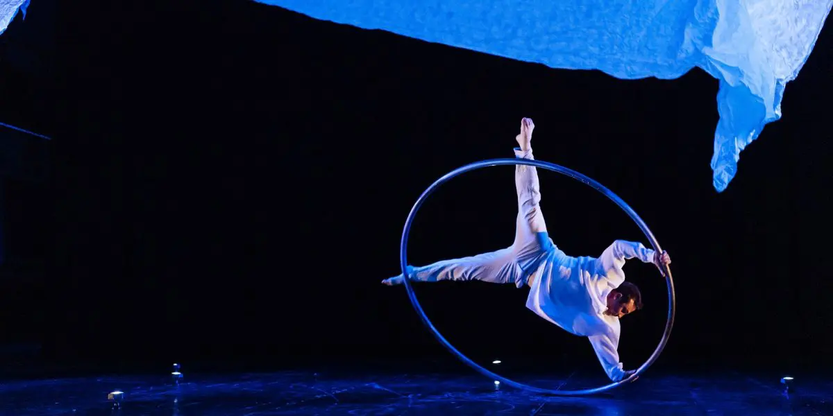 Un interprète en costume blanc, rappelant le style de Robert Cookson, se tient en équilibre horizontal à l'intérieur d'un grand anneau métallique sur une scène sombre, éclairée par une lumière bleue et un tissu drapé au-dessus de sa tête.