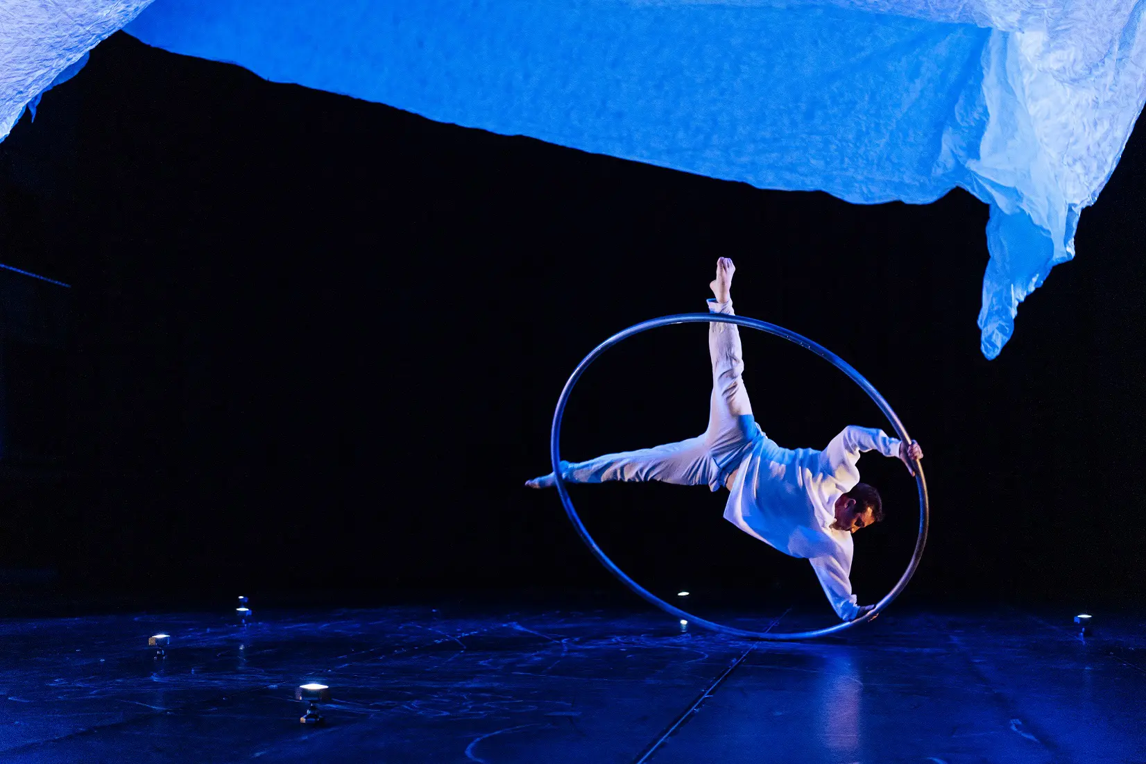 Un interprète en costume blanc, rappelant le style de Robert Cookson, se tient en équilibre horizontal à l'intérieur d'un grand anneau métallique sur une scène sombre, éclairée par une lumière bleue et un tissu drapé au-dessus de sa tête.