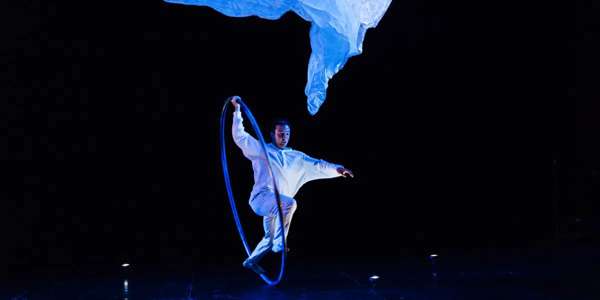 Un artiste vêtu de blanc, rappelant l'art de Robert Cookson, se tient en équilibre sur un pied à l'intérieur d'un grand cerceau sur une scène sombre, avec un éclairage bleu dramatique et un tissu blanc flottant au-dessus de sa tête.