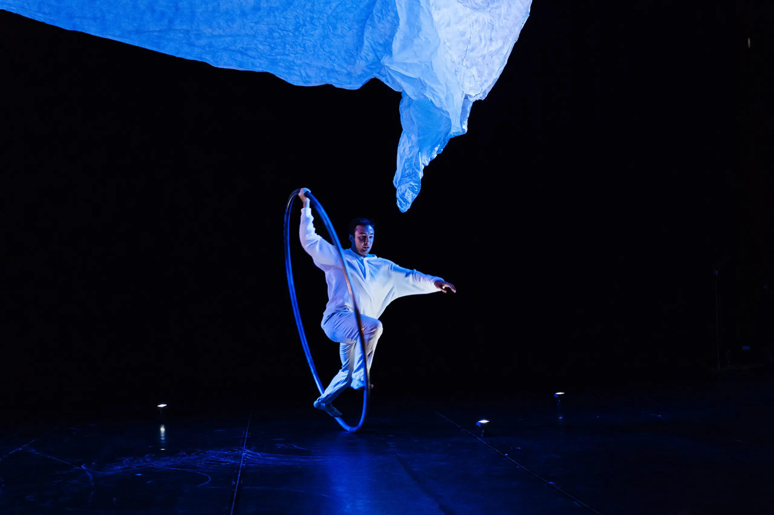 Un artiste vêtu de blanc, rappelant l'art de Robert Cookson, se tient en équilibre sur un pied à l'intérieur d'un grand cerceau sur une scène sombre, avec un éclairage bleu dramatique et un tissu blanc flottant au-dessus de sa tête.