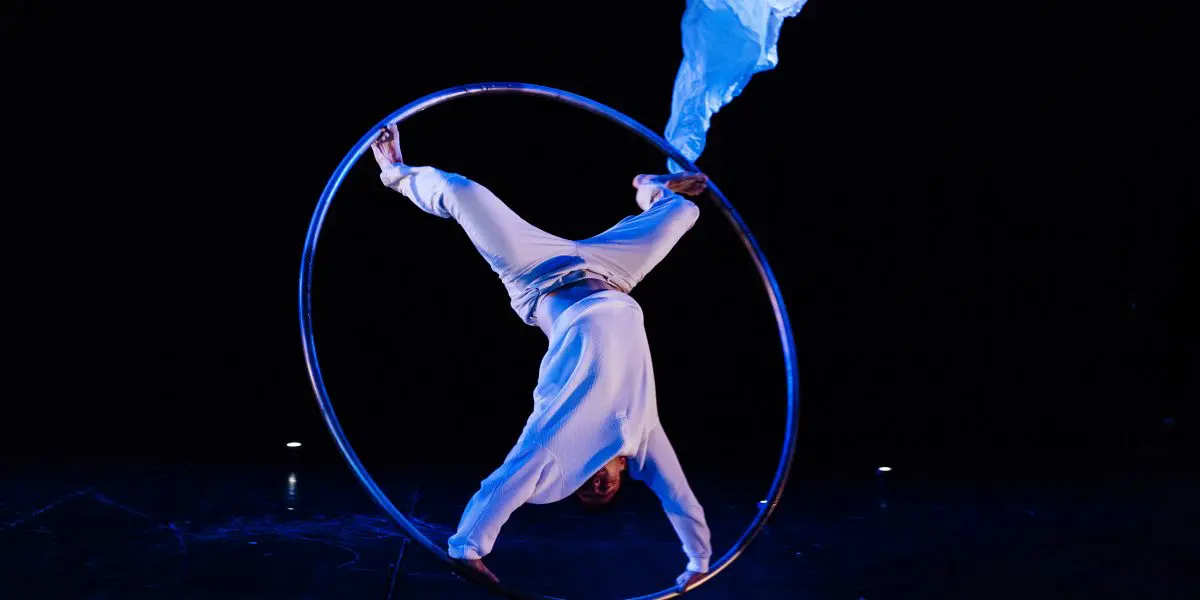 Un artiste en blanc, rappelant l'art de Robert Cookson, se tient en équilibre à l'intérieur d'un grand cerceau métallique sur une scène sombre, surplombée d'un tissu bleu fluide.