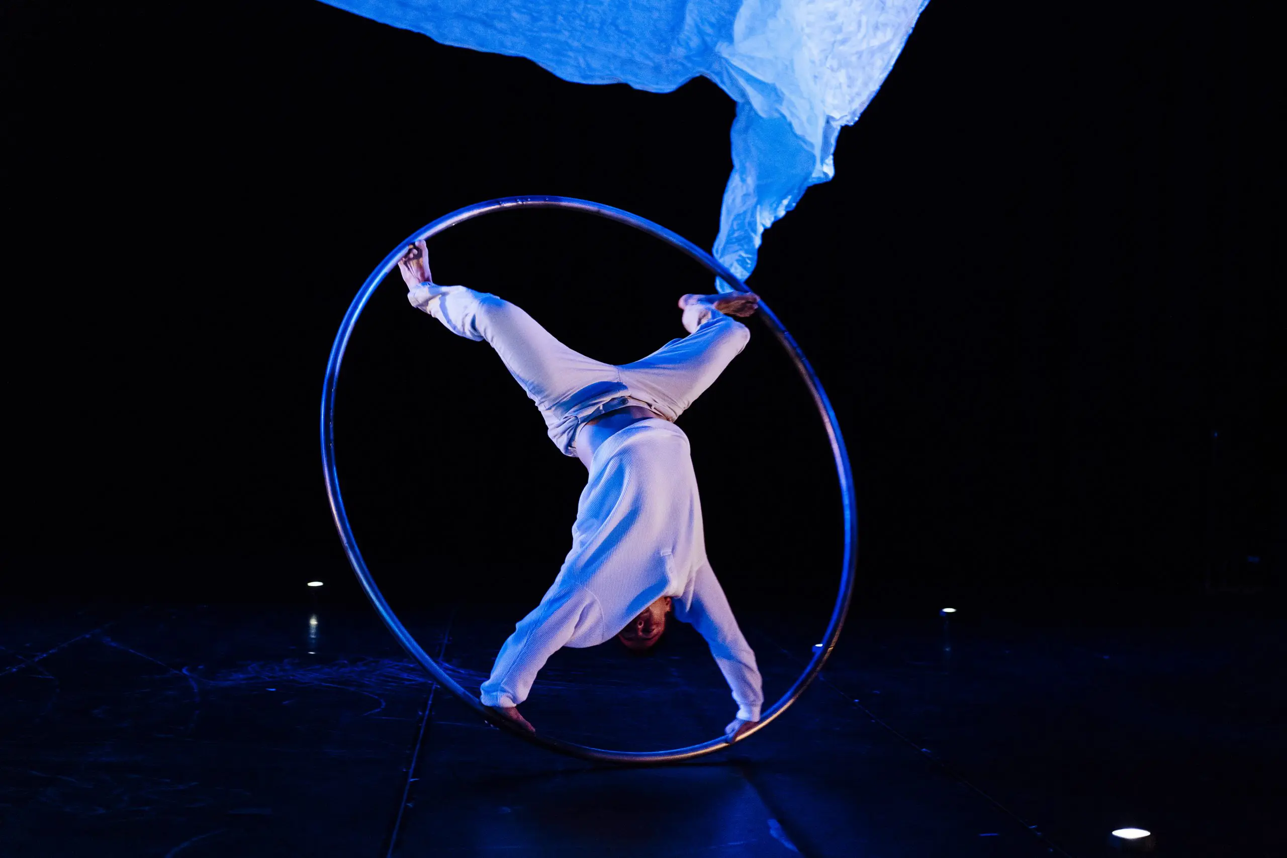 Un artiste en blanc, rappelant l'art de Robert Cookson, se tient en équilibre à l'intérieur d'un grand cerceau métallique sur une scène sombre, surplombée d'un tissu bleu fluide.