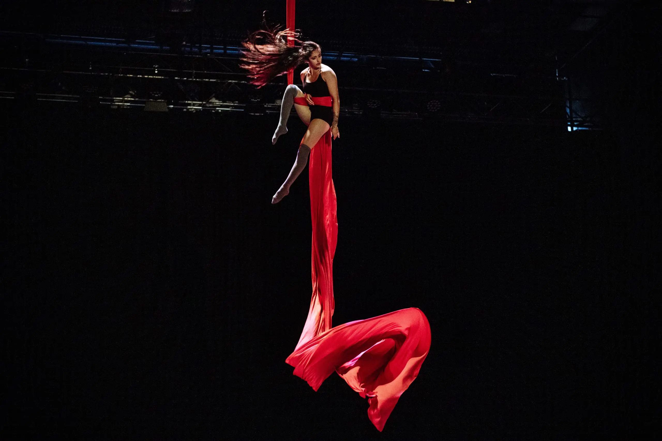 Une voltigeuse, Leela Masuret, se produit en hauteur, suspendue à des soies rouges fluides sur un fond sombre, ses cheveux et le tissu étant saisis en mouvement de manière spectaculaire.