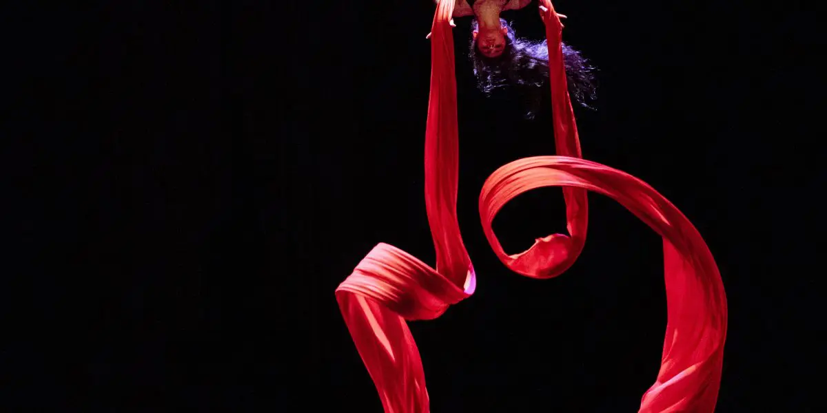 L'artiste aérienne Leela Masuret est suspendue à l'envers à des rubans de soie rouge, le tissu fluide se tordant autour d'elle, sur un fond sombre.