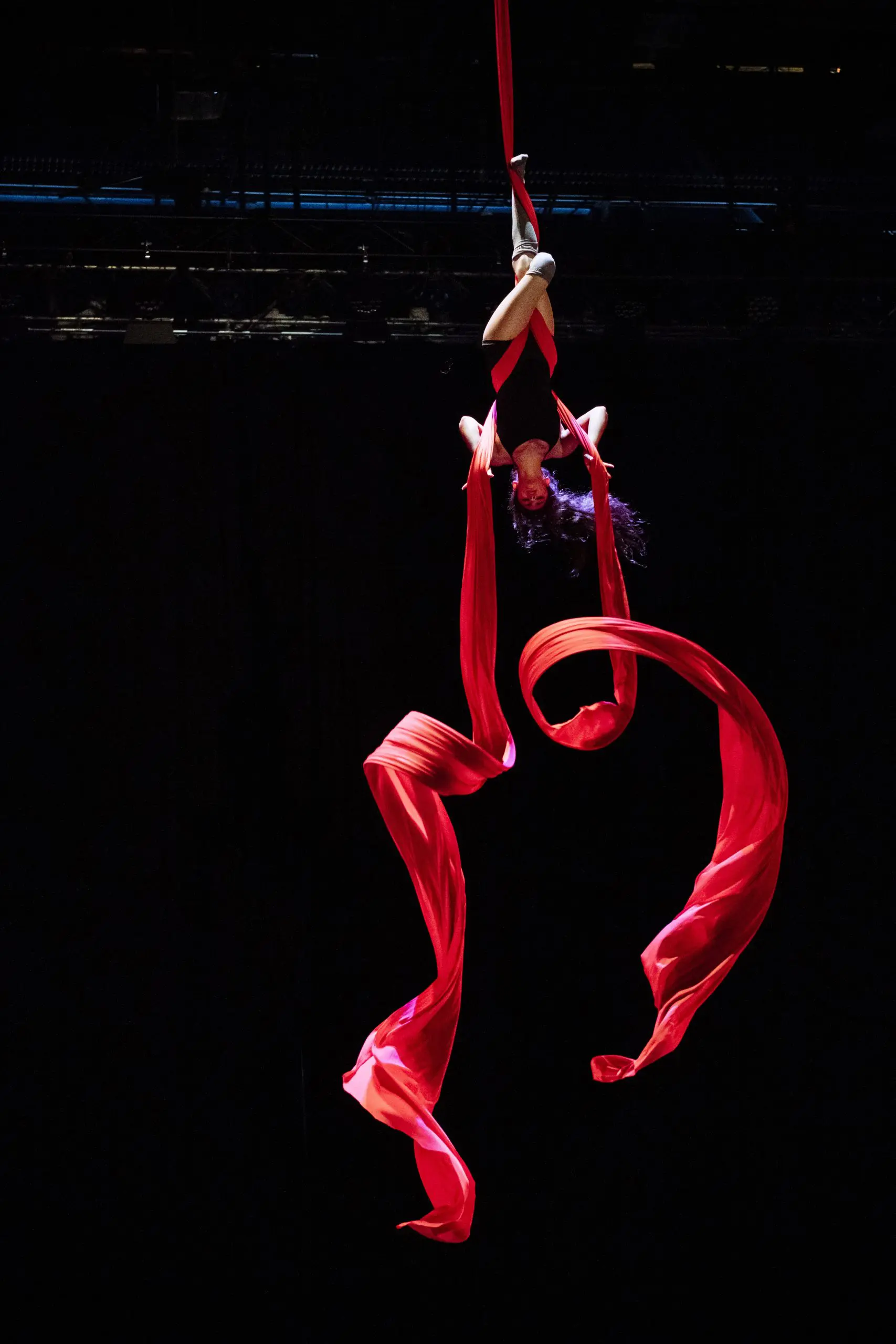 L'artiste aérienne Leela Masuret est suspendue à l'envers à des rubans de soie rouge, le tissu fluide se tordant autour d'elle, sur un fond sombre.