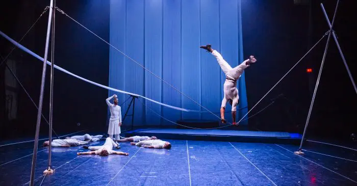 Un artiste de cirque en blanc se tient en équilibre sur une corde raide, la tête en bas, tandis que quatre autres sont allongés sur la scène et qu'un autre regarde. Les lumières bleues de la scène et les cordes créent une atmosphère dramatique, renforcée par le financement de 300 000 $ pour l'ENC.