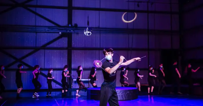 Un artiste masqué souffle une bulle avec une baguette sur scène pendant les présentations de fin d'année des élèves du programme Cirque-études secondaires, tandis que des danseurs masqués vêtus de noir se déplacent énergiquement sous un éclairage violet-bleu.