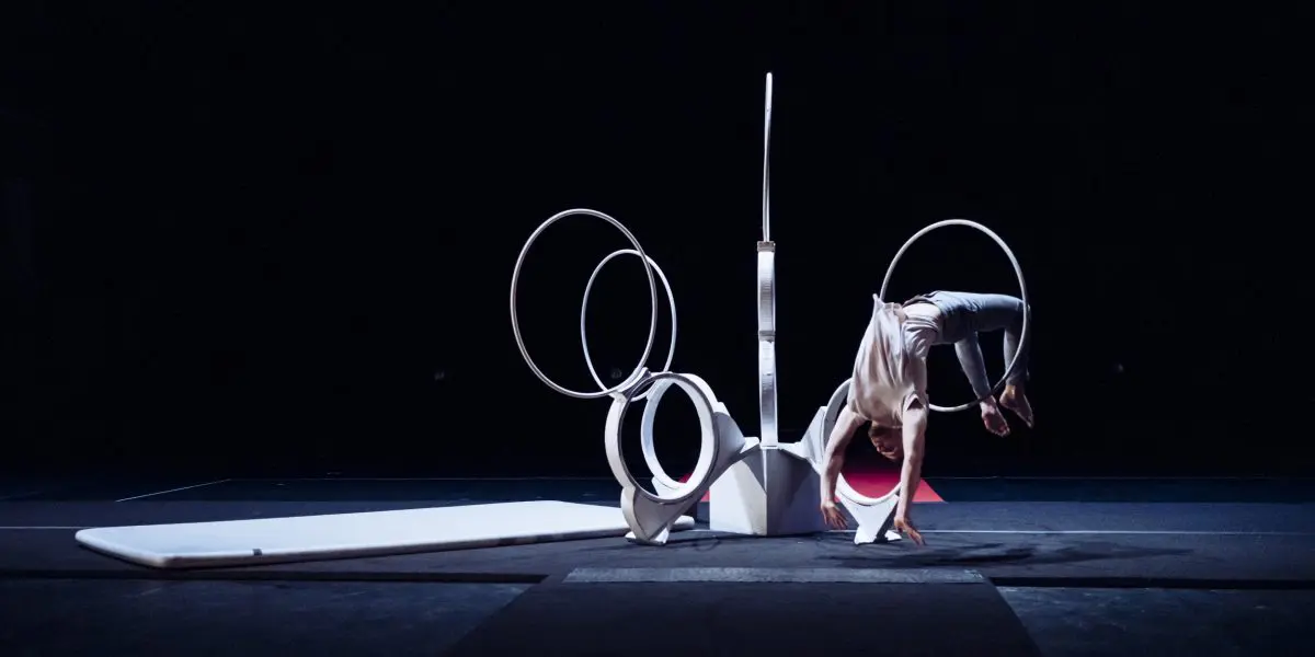 Laurent Racicot se tient en équilibre la tête en bas, les jambes pliées, à travers de grands anneaux blancs sur une scène faiblement éclairée, entourée de tapis de gymnastique et d'accessoires géométriques.