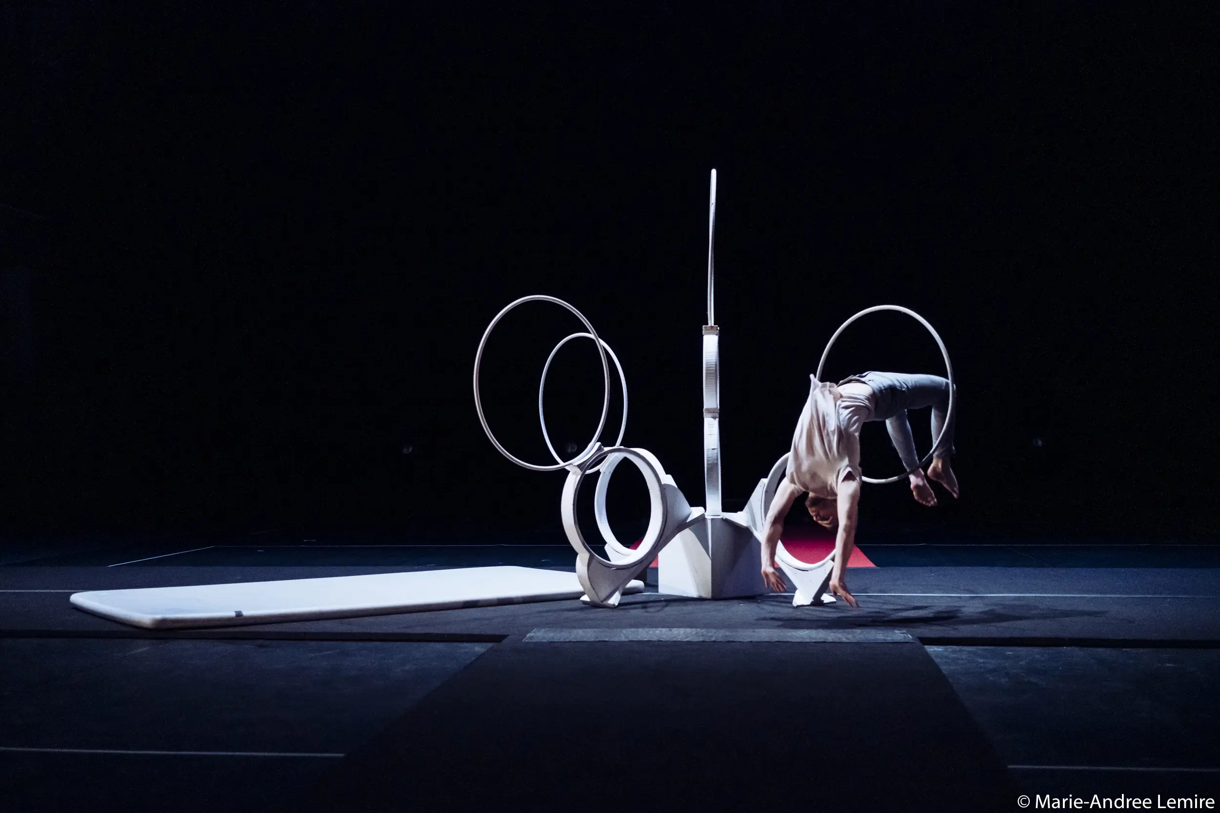Laurent Racicot se tient en équilibre la tête en bas, les jambes pliées, à travers de grands anneaux blancs sur une scène faiblement éclairée, entourée de tapis de gymnastique et d'accessoires géométriques.
