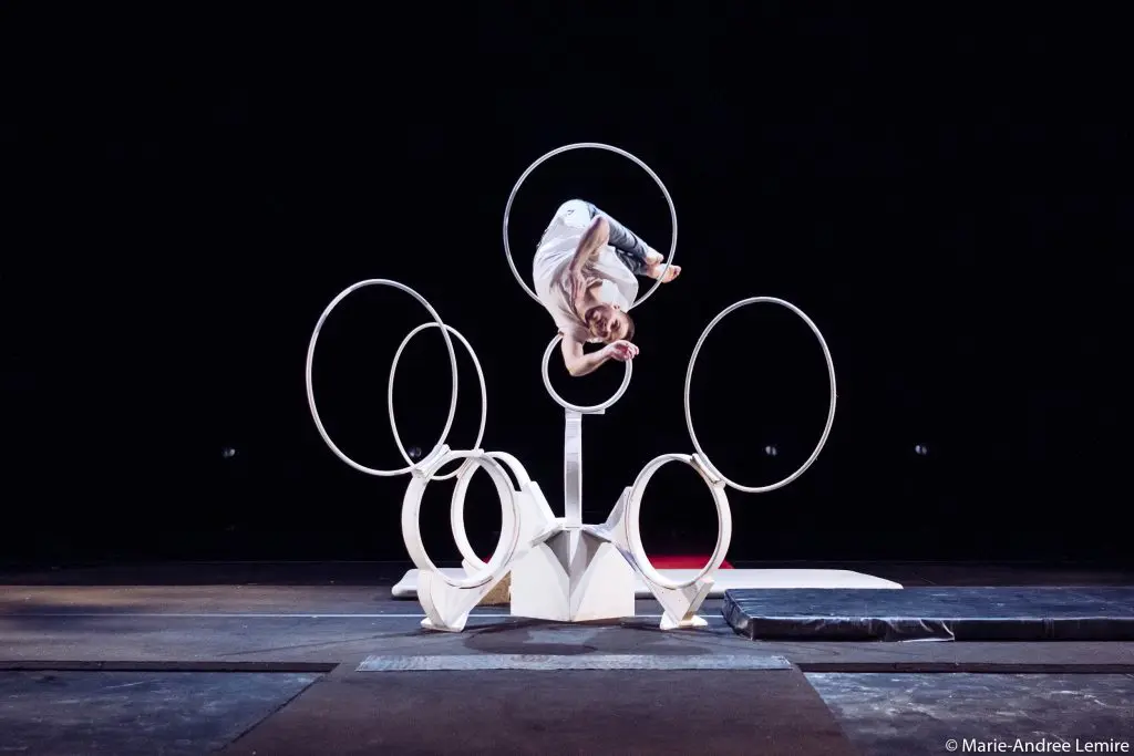 Laurent Racicot, vêtu de blanc, se tient en équilibre à l'intérieur d'un grand anneau circulaire. Plusieurs autres anneaux sont montés sur une structure blanche sur la scène, sur un fond sombre.