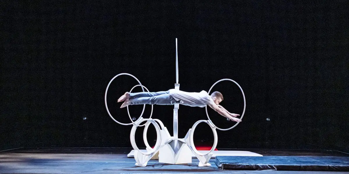 Laurent Racicot, vêtu de blanc, se tient en équilibre horizontal en saisissant deux grands anneaux circulaires montés sur une structure blanche à anneaux multiples, sur un fond de scène sombre.