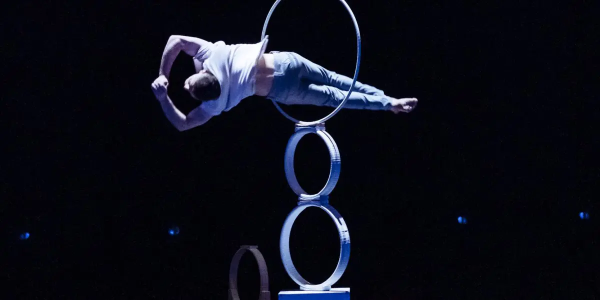 Laurent Racicot se tient en équilibre horizontal sur un bras au sommet d'une structure circulaire composée de trois anneaux empilés verticalement, attirant l'attention sur une scène faiblement éclairée.