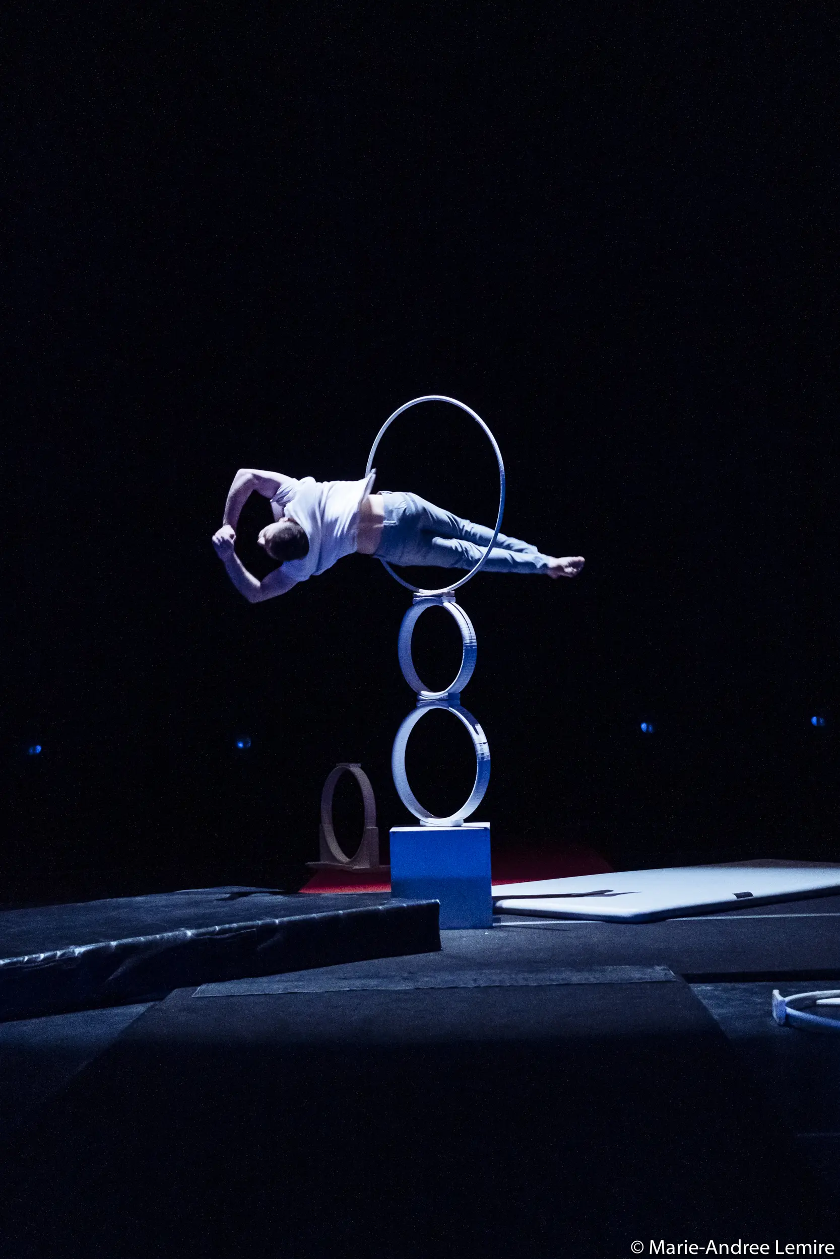 Laurent Racicot se tient en équilibre horizontal sur un bras au sommet d'une structure circulaire composée de trois anneaux empilés verticalement, attirant l'attention sur une scène faiblement éclairée.