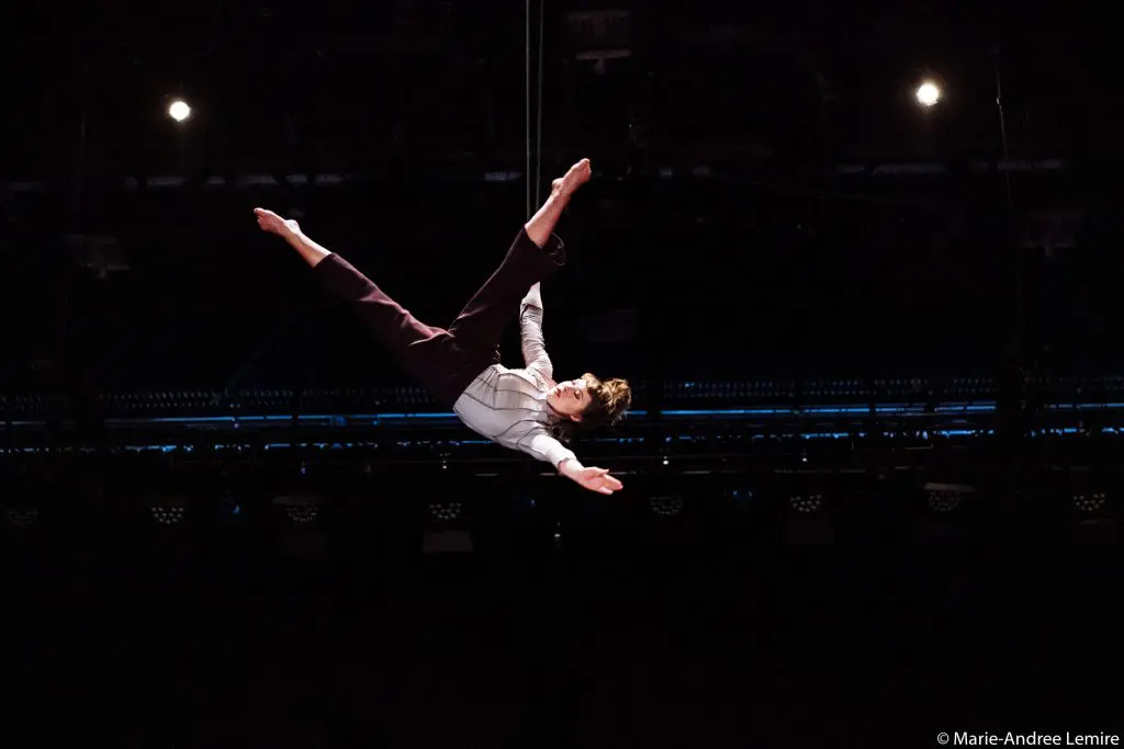 Mathilde Richer, performeuse suspendue dans les airs par une corde, forme un X avec les bras et les jambes tendus sur un fond sombre, vêtue d'un pantalon sombre et d'une chemise claire.