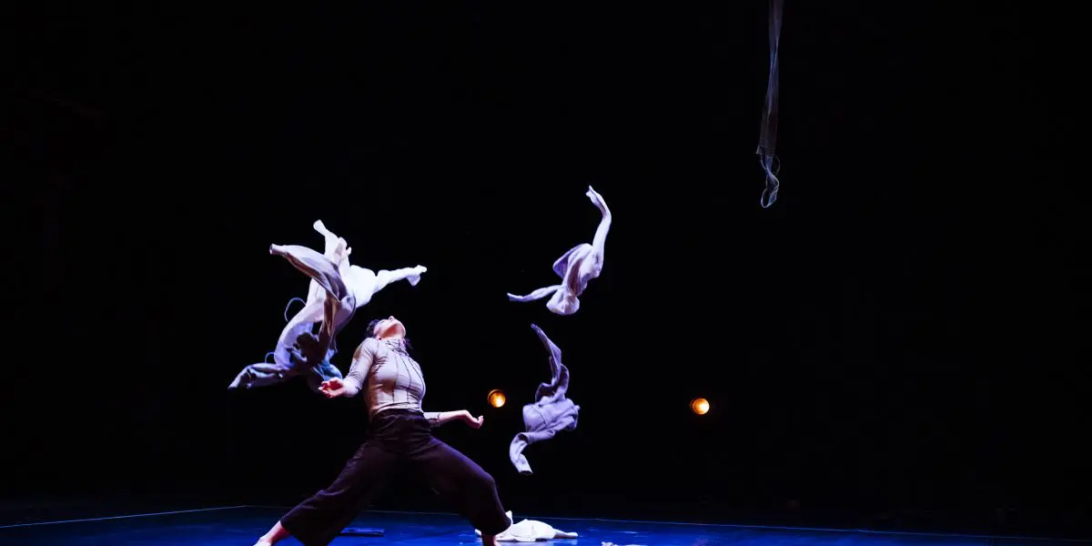 Mathilde Richer se tient sur une scène sombre, en plein mouvement, lançant des vêtements en l'air. Les vêtements sont suspendus au-dessus d'elle, tandis qu'elle se penche en arrière, les bras levés, éclairée par des lumières de scène rougeoyantes en arrière-plan.
