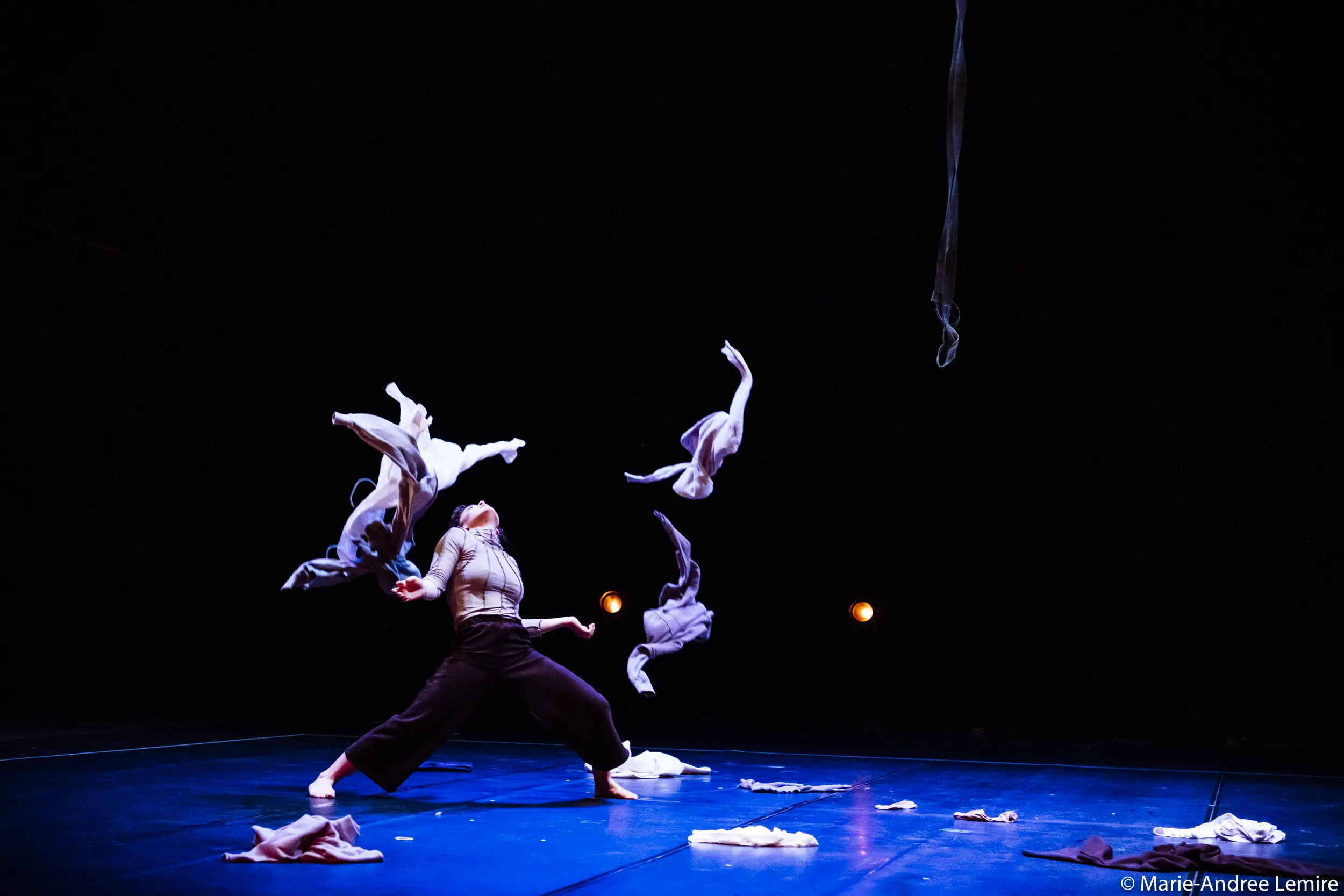 Mathilde Richer se tient sur une scène sombre, en plein mouvement, lançant des vêtements en l'air. Les vêtements sont suspendus au-dessus d'elle, tandis qu'elle se penche en arrière, les bras levés, éclairée par des lumières de scène rougeoyantes en arrière-plan.