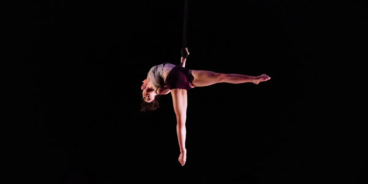 L'artiste aérienne Mathilde Richer se produit suspendue dans les airs, tenant une sangle d'un bras, le corps tendu à l'horizontale sur un fond sombre.