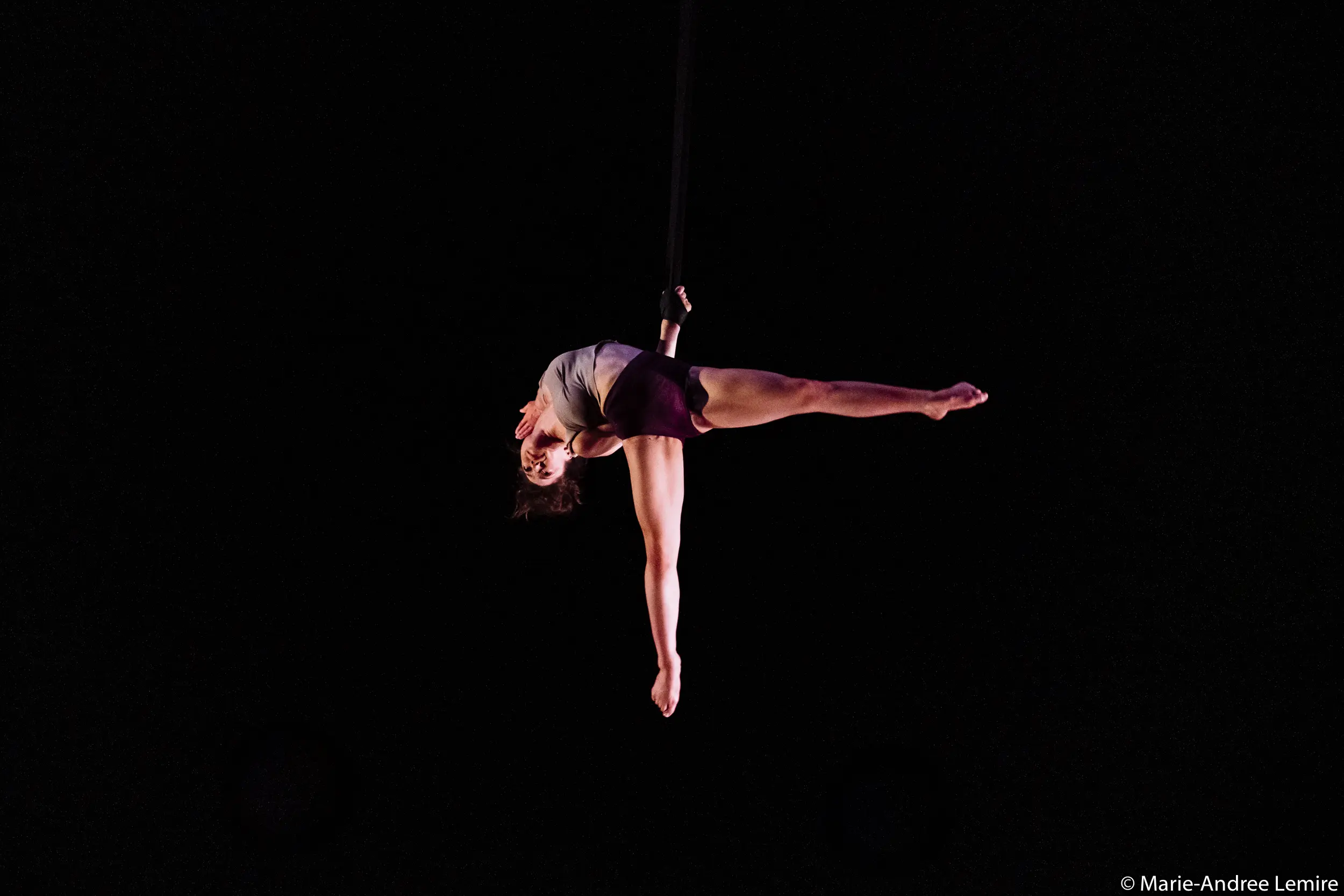 L'artiste aérienne Mathilde Richer se produit suspendue dans les airs, tenant une sangle d'un bras, le corps tendu à l'horizontale sur un fond sombre.