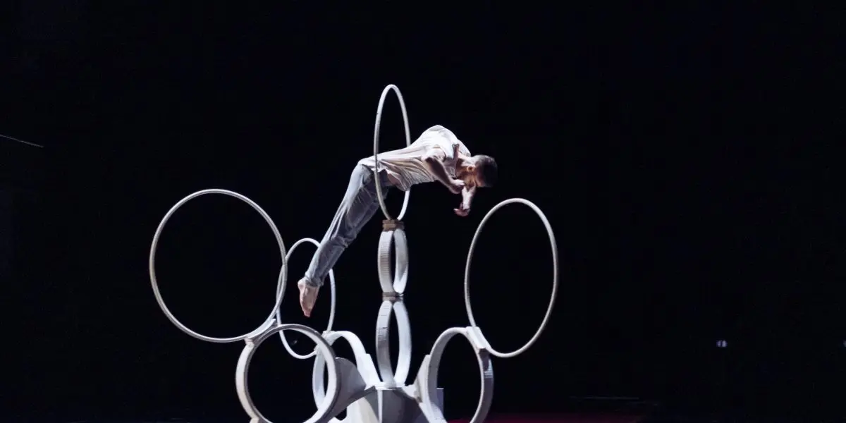 Un interprète, qui rappelle Laurent Racicot, se tient en équilibre horizontal sur de grands cerceaux imbriqués, disposés de manière sculpturale sur une scène faiblement éclairée.
