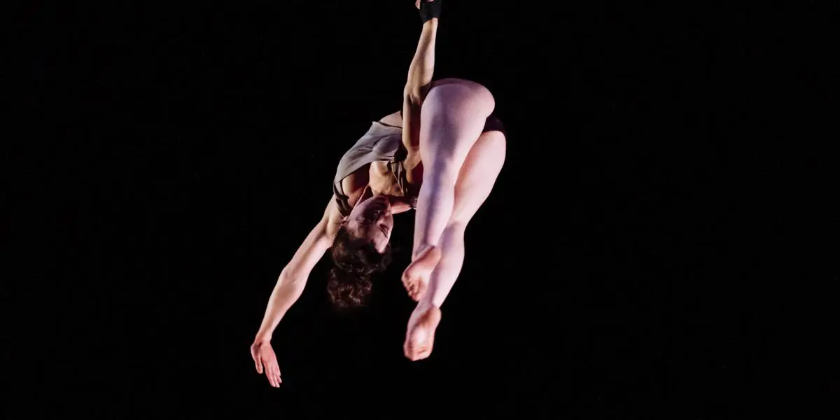 L'artiste aérienne Mathilde Richer, vêtue d'un justaucorps, est suspendue dans les airs, arquant gracieusement son dos et étendant ses bras et ses jambes sur un fond noir.