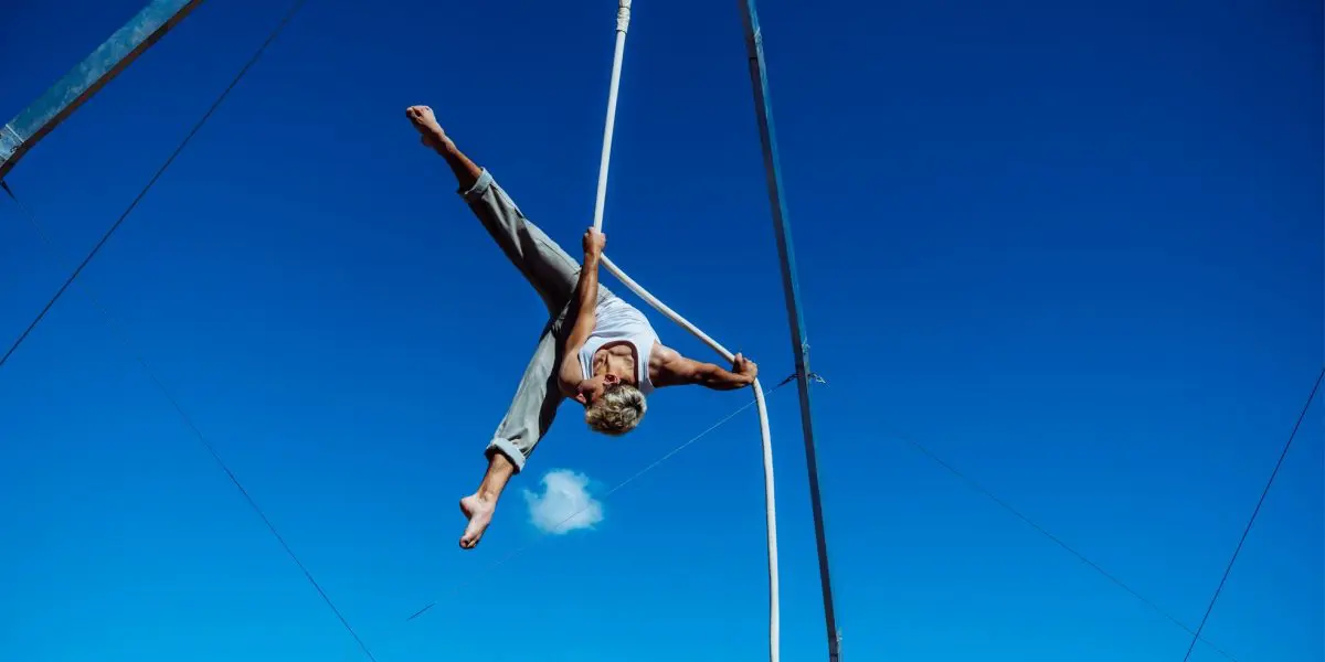 Une personne effectue une pose acrobatique aérienne sur des soies blanches, suspendue en hauteur dans un ciel bleu clair, avec des structures de soutien métalliques visibles - un spectacle extérieur à couper le souffle 2021 entre-temps des étudiant-e-s de l'ENC.