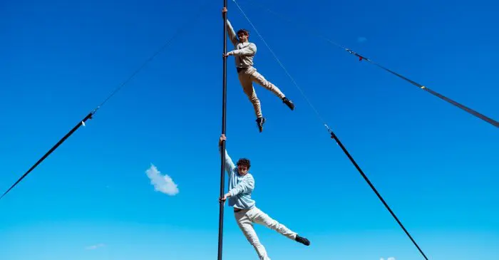 Deux personnes en vêtements clairs font des acrobaties sur un grand mât sous un ciel bleu clair pendant le spectacle extérieur 2021 entre-temps des étudiant-e-s de l'ENC, avec des cordes ancrant le mât et des arbres à l'arrière-plan.