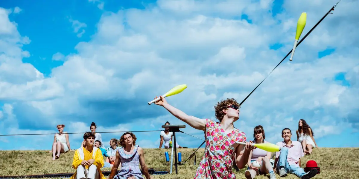 Une personne vêtue d'une robe à fleurs jongle avec des massues en plein air sur une colline herbeuse lors du spectacle extérieur 2021 entre-temps des étudiant-e-s de l'ENC, alors que plusieurs personnes sont assises et regardent sous un ciel bleu avec des nuages épars.