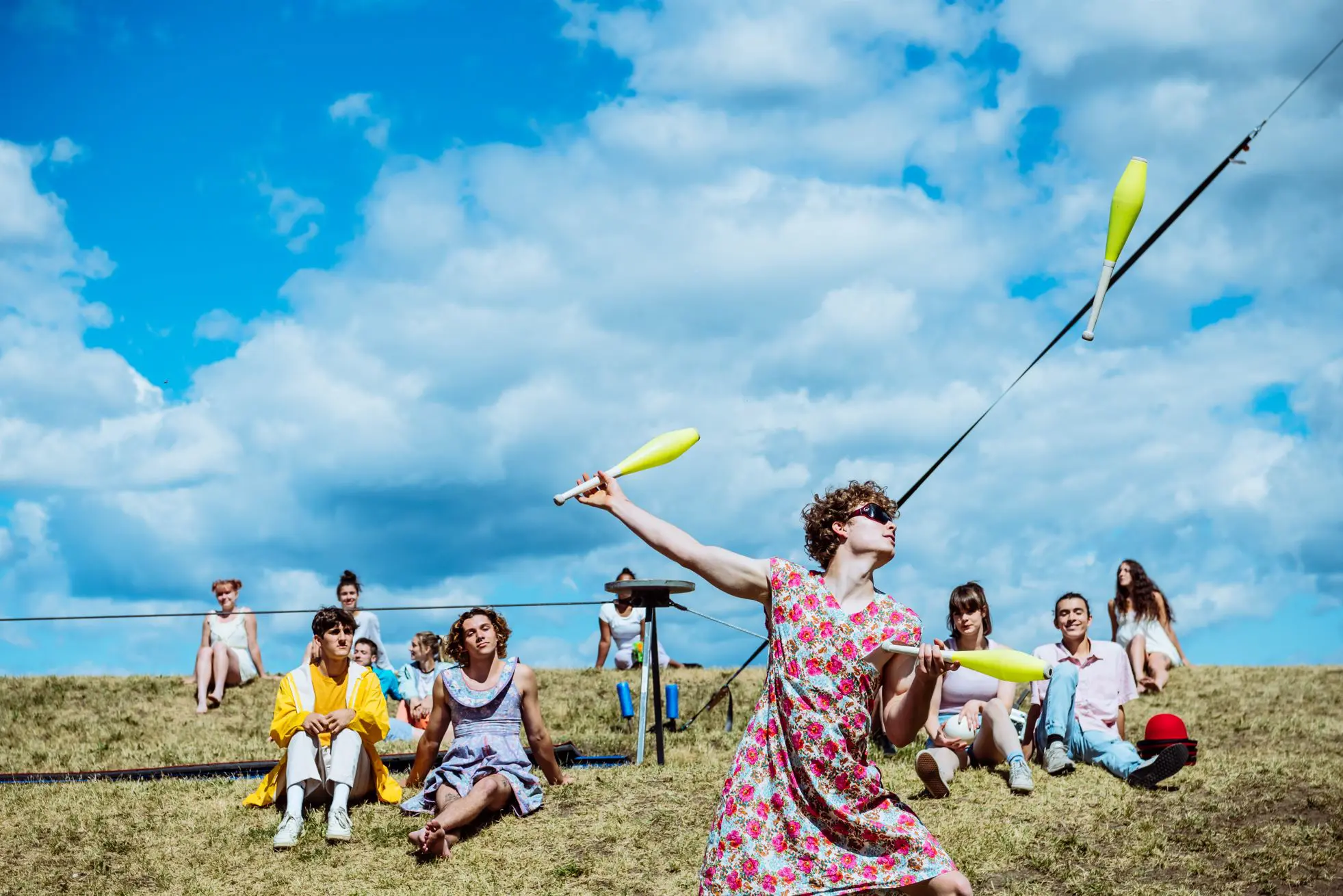 Une personne vêtue d'une robe à fleurs jongle avec des massues en plein air sur une colline herbeuse lors du spectacle extérieur 2021 entre-temps des étudiant-e-s de l'ENC, alors que plusieurs personnes sont assises et regardent sous un ciel bleu avec des nuages épars.