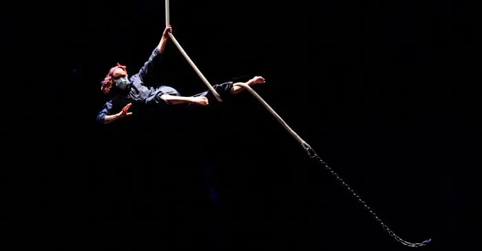 Une personne portant une tenue sombre et un bandeau rouge exécute un numéro d'acrobatie aérienne en s'agrippant à un poteau suspendu au-dessus de la scène sombre pendant Triptyque 2021, le spectacle des étudiant-e-s de la 2e année de l'ENC.
