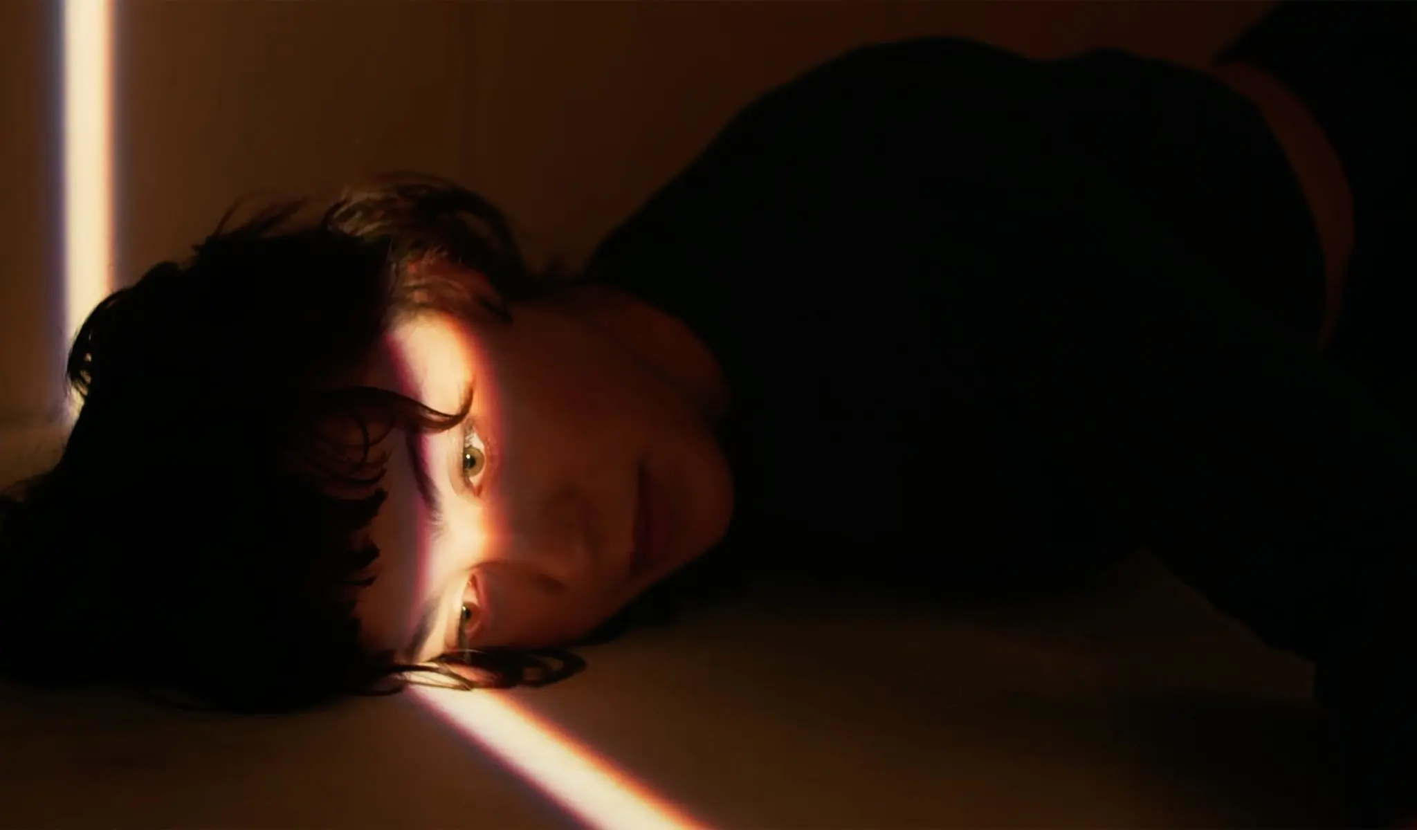 Une personne aux cheveux courts et noirs est allongée sur le côté dans une pièce faiblement éclairée, un étroit faisceau de lumière illuminant un œil, évoquant l'ambiance dramatique de *le film des finissant-e-s 2021 la somme de nos rêves*.